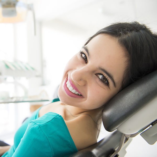 a lady with a black hair  sitting on a dental chair looking at her left showing her beautiful smile