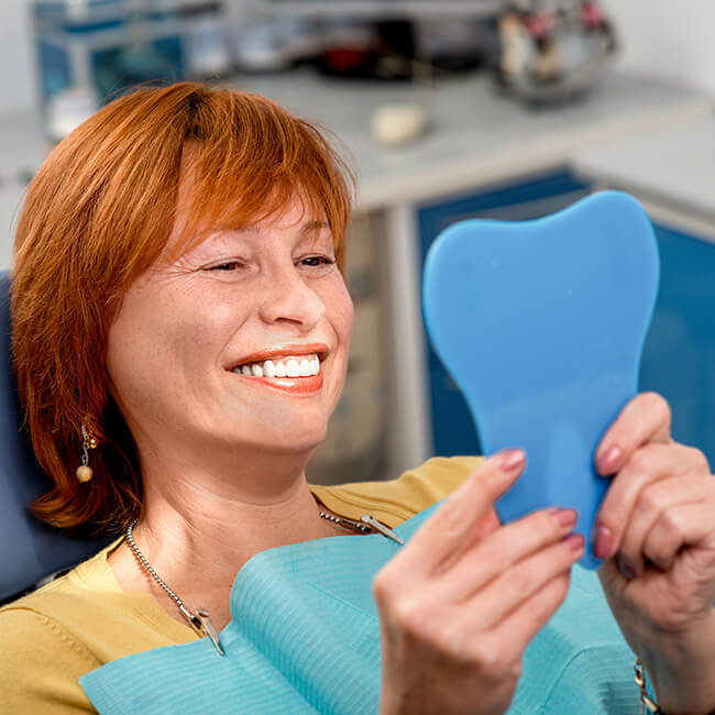 A woman lying on a dental chair is while looking at her smile in the mirror she is holding