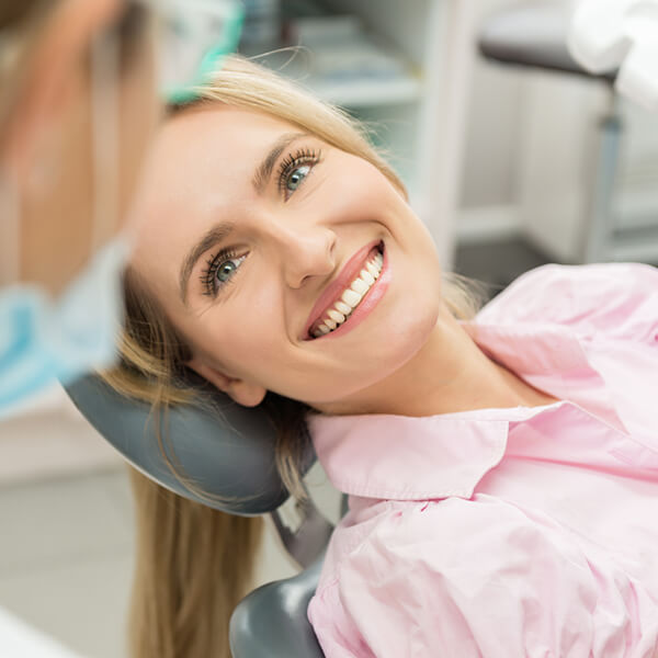 A lady with blonde hair wearing a pink blouse sitting in dental chair while looking at the Doctor